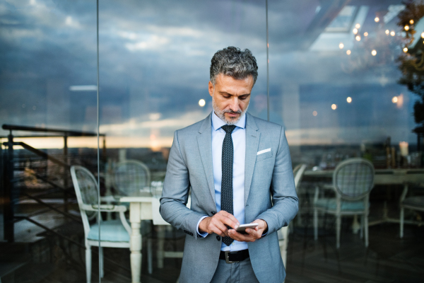 Mature businessman with smartphone standing in an outdoor hotel cafe. Man text messaging.