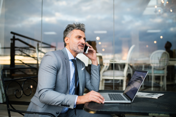 Mature businessman with laptop and smartphone sitting in an outdoor hotel cafe. Man making a phone call.