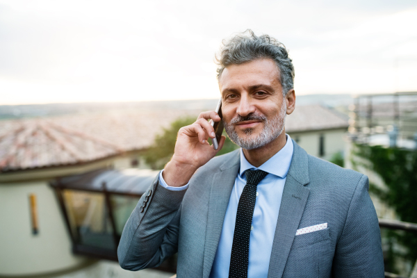 Mature businessman with smartphone standing in an outdoor hotel cafe. Man making a phone call.