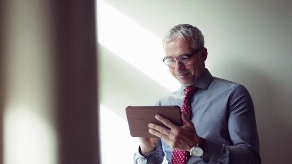 A mature confident businessman on a business trip standing in a hotel room, using tablet.