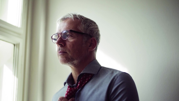 Mature businessman with glasses on a business trip standing in a hotel room, getting dressed. Slow motion.