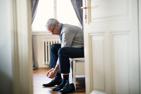 Mature businessman with glasses on a business trip sitting ina hotel room, tying shoelaces.