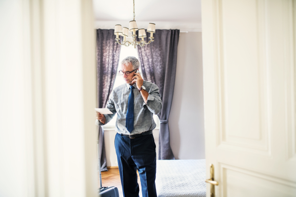 Mature businessman with glasses on a business trip standing in a hotel room, making phone call.