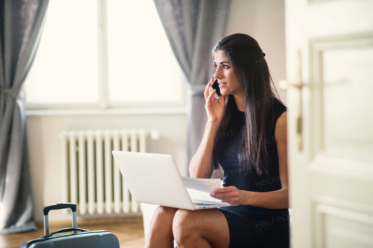 Young businesswoman with suitcase on a business trip sitting in a hotel room, using smartphone and laptop.