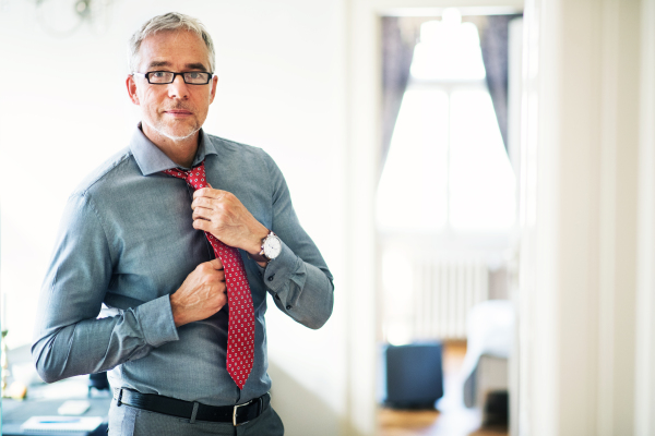 Mature businessman with glasses on a business trip standing in a hotel room, getting dressed. Copy space.