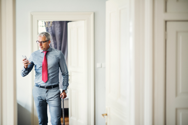 Mature businessman with glasses and suitcase on a business trip standing in a hotel room, using smartphone. Copy space.