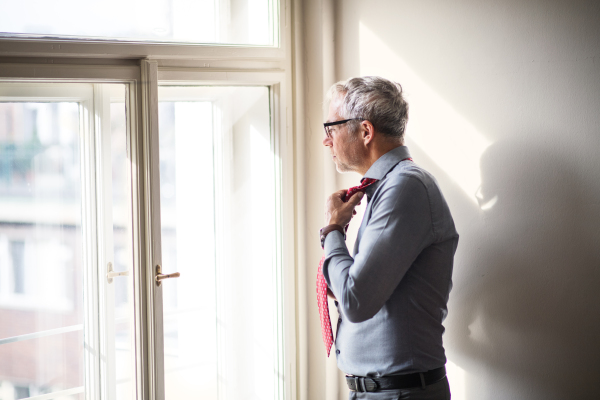 Mature businessman with glasses on a business trip standing in a hotel room, looking out of a window when getting dressed.