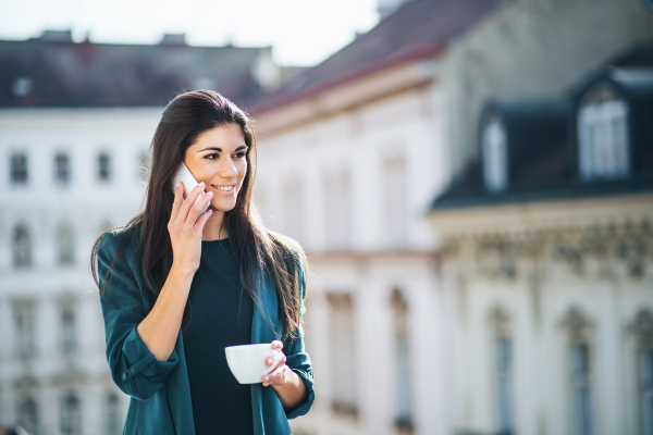 Young businesswoman with coffee and smartphone standing on a terrace outside an office in city, making a phone call. Copy space.