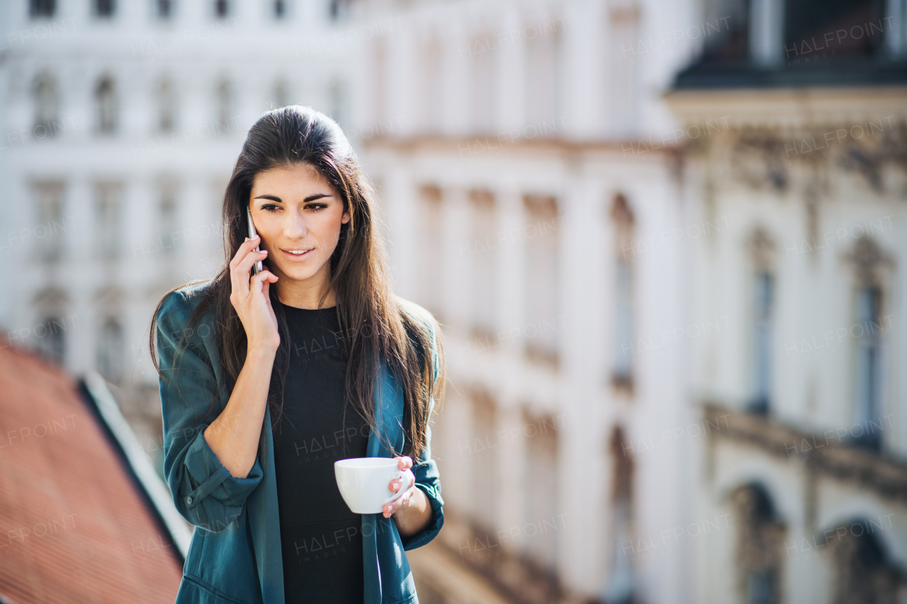 Young businesswoman with coffee and smartphone standing on a terrace outside an office in city, making a phone call. Copy space.