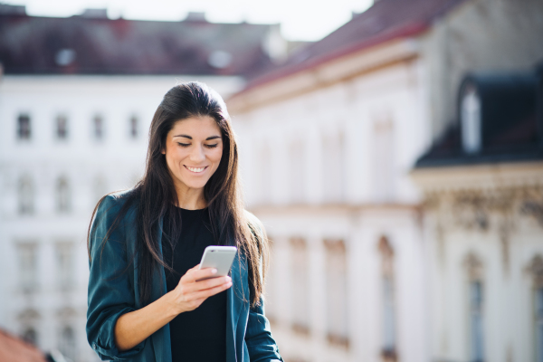 Young businesswoman with smartphone standing on a terrace outside an office in city. Copy space.