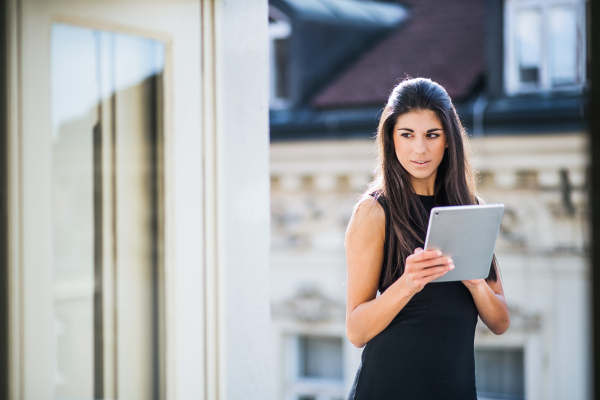 Young businesswoman with tablet standing on a terrace outside an office in city. Copy space.