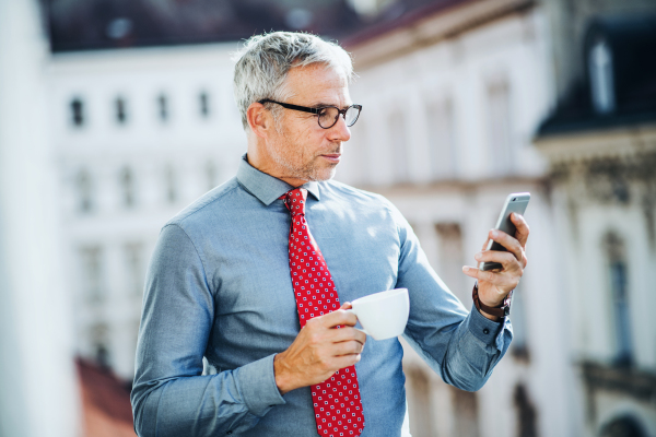 A mature businessman with coffee and smartphone standing on a terrace in an office in city, texting.