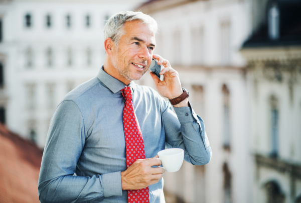 A mature businessman with smartphone and coffee standing on a terrace in an office in city, making a phone call.