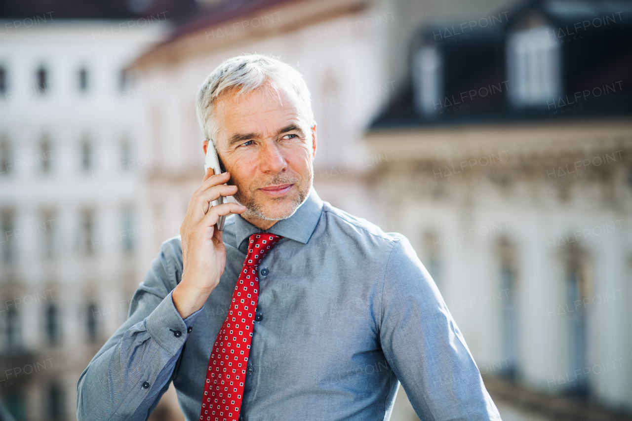 A mature businessman with smartphone standing on a terrace in an office in city, making a phone call.