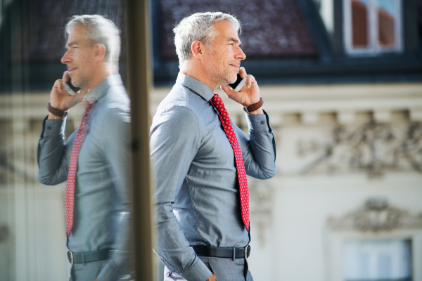 A mature businessman with smartphone standing on a terrace in an office in city, making a phone call.