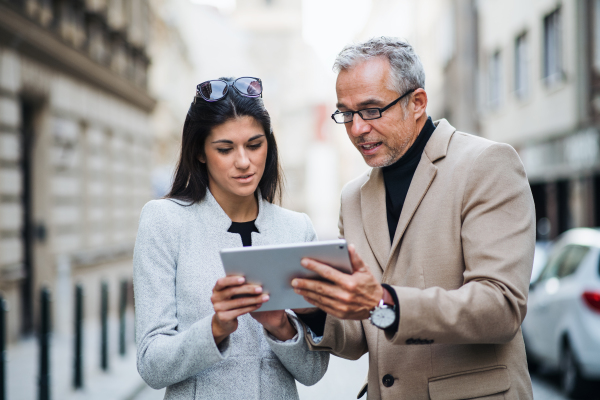 Mature man and young woman business partners with tablet standing outdoors in city of Prague, talking.