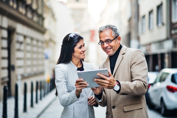 Mature man and young woman business partners with tablet standing outdoors in city of Prague, talking.