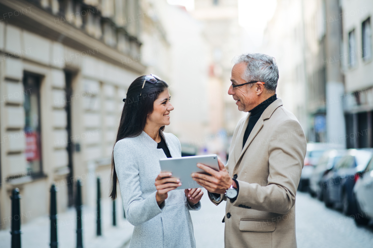 Mature man and young woman business partners with tablet standing outdoors in city of Prague, talking.