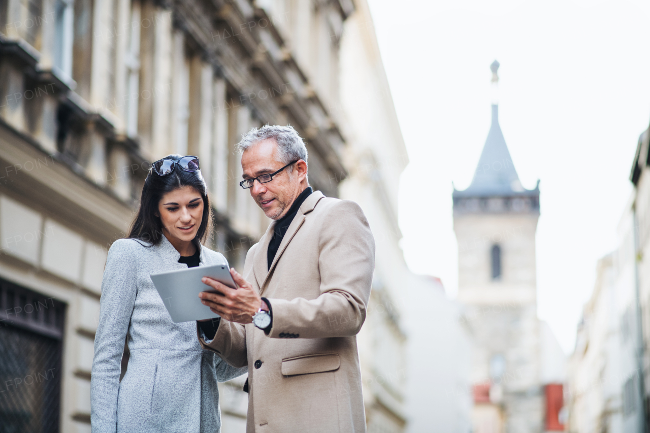 Mature man and young woman business partners with tablet standing outdoors in city of Prague, talking.