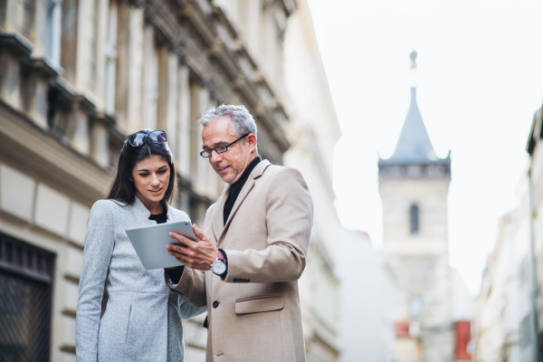 Mature man and young woman business partners with tablet standing outdoors in city of Prague, talking.