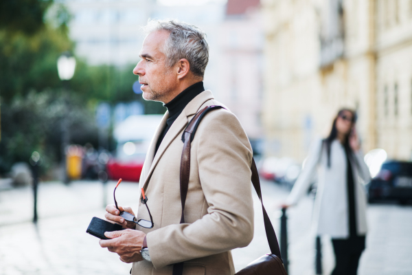 Mature businessman standing on a street in city, holding glasses. Copy space.