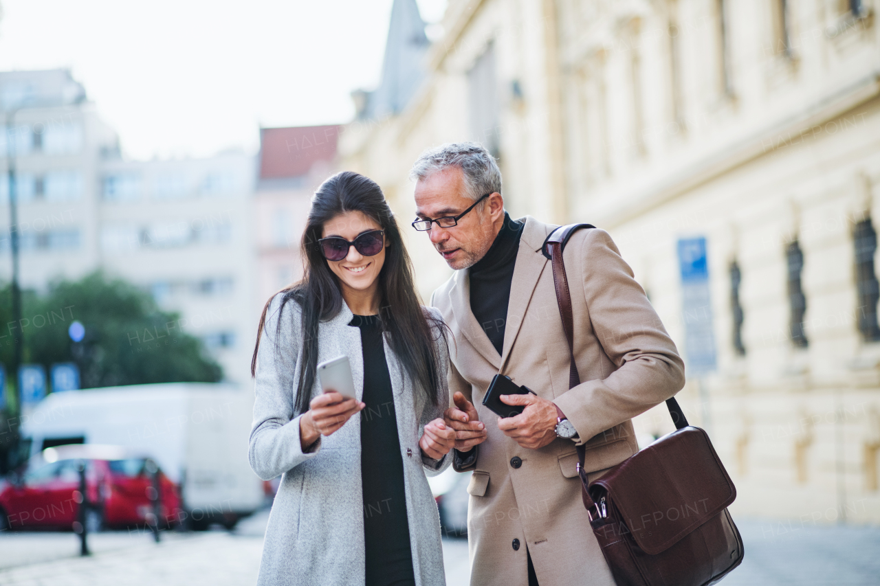 Mature man and young woman business partners with smartphone standing outdoors in city of Prague, talking.