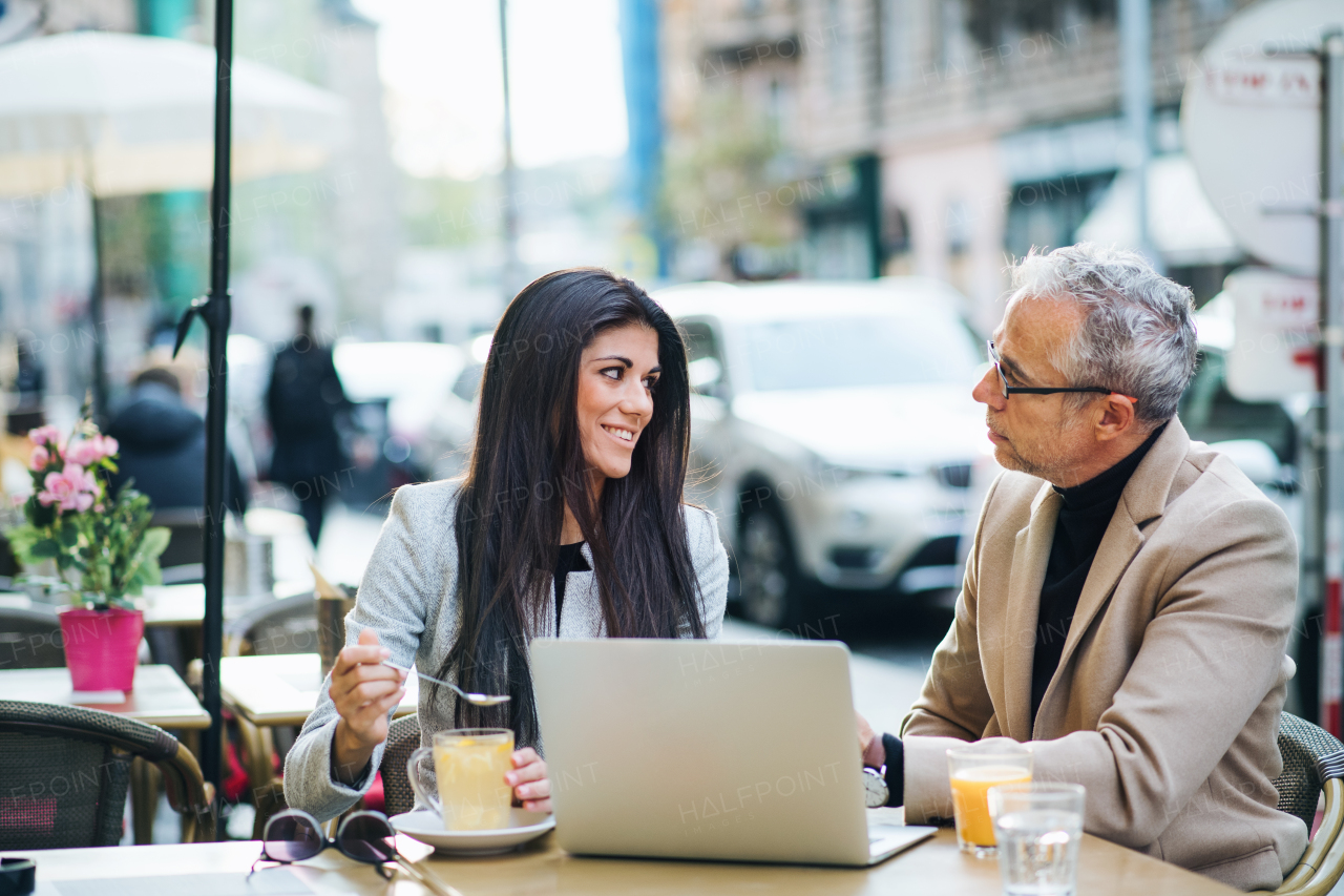 Man and woman business partners with laptop and drinks sitting in a cafe in city, talking.