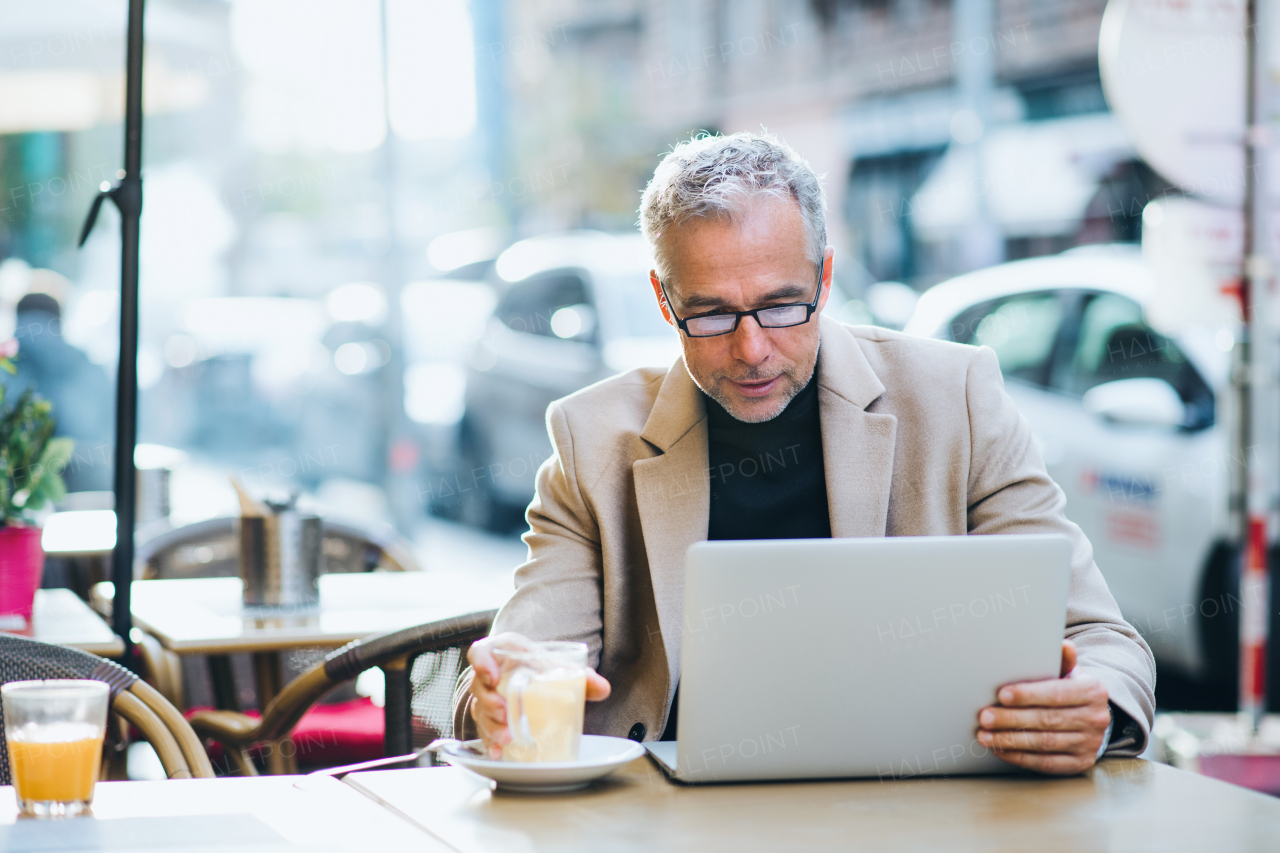 Mature businessman with laptop sitting in a cafe in city, working.