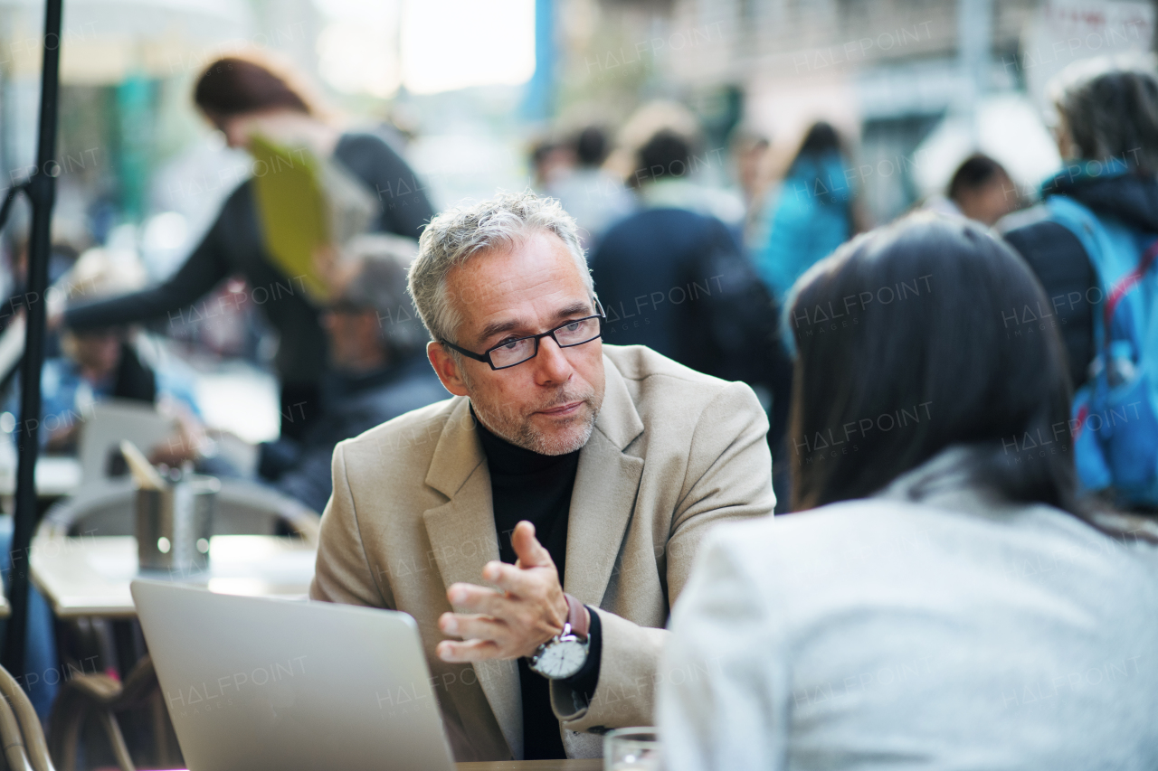 Man and unrecognizable woman business partners with laptop sitting in a crowded cafe in city, talking.