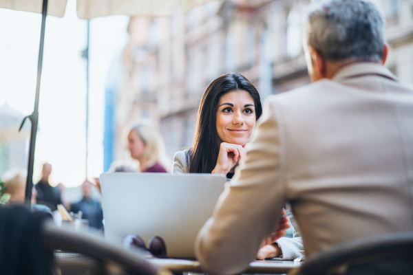 Man and unrecognizable woman business partners with laptop sitting in a crowded cafe in city, talking.