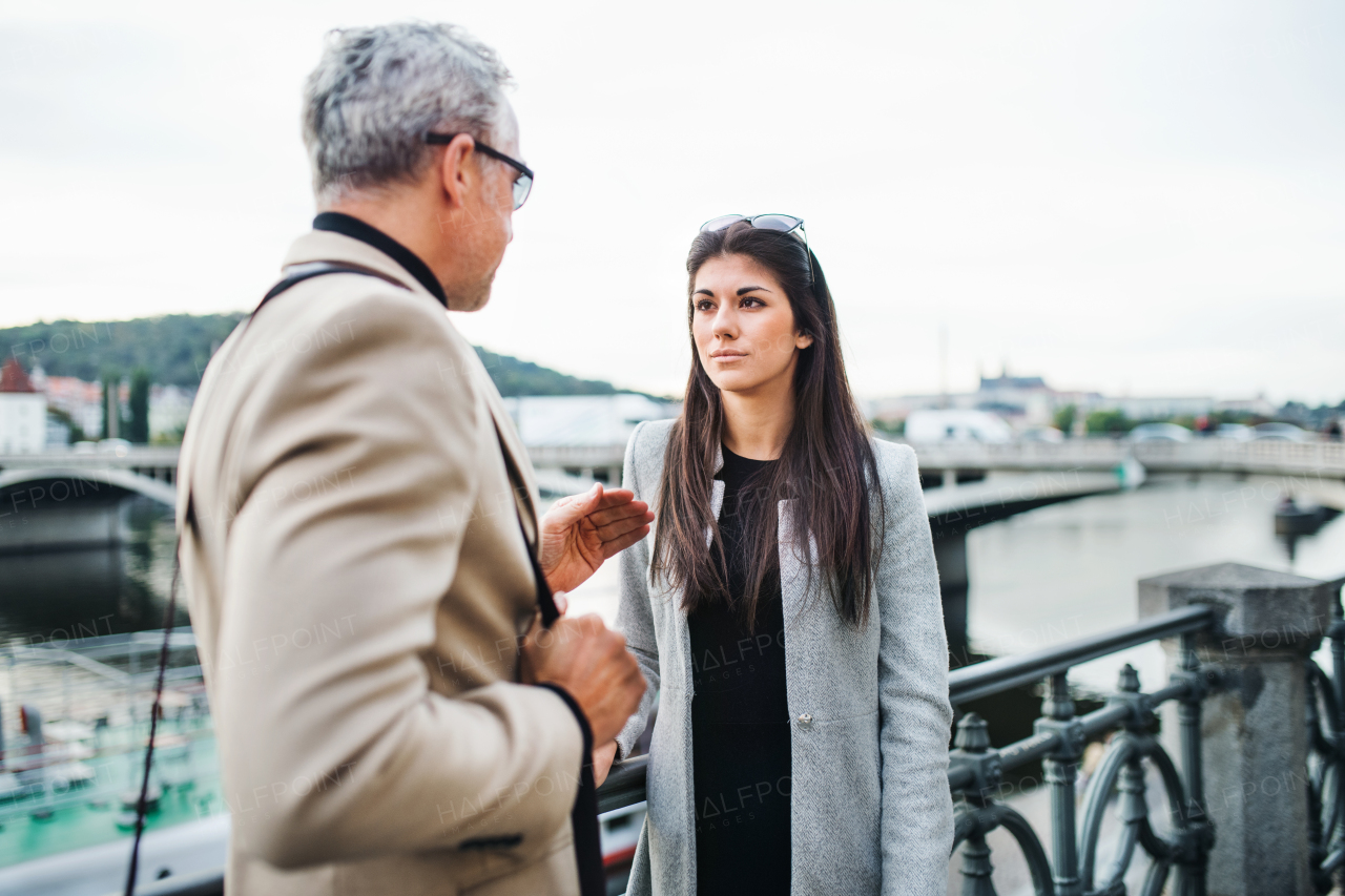 Mature man and young woman business partners walking by a river in city of Prague, talking.