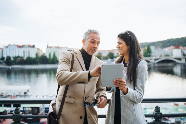 Man and woman business partners with tablet standing by a river in city of Prague, talking.