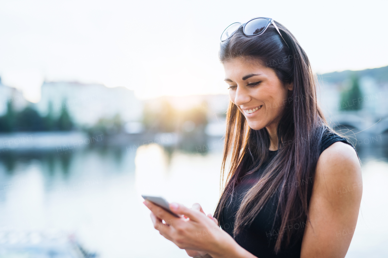 Beautiful woman in black dress with sunglasses and smartphone standing by a river in city of Prague, text messaging.