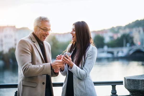 Man and woman business partners standing by a river in city at sunset, looking at smartphone and expressing excitement.