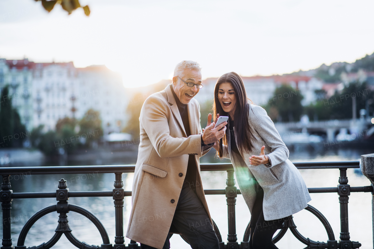 Man and woman business partners standing by a river in city at sunset, looking at smartphone and expressing excitement.