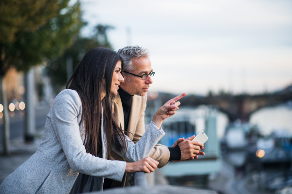 Man and woman business partners standing by a river in city of Prague, pointing to something.