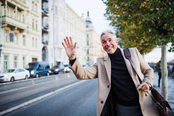 Mature businessman with bag standing on the street in city of Prague, raising his hand to hail a taxi cab.