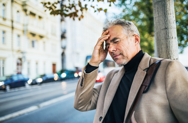 Mature businessman in pain standing on a street in city, holding forehead. Copy space.