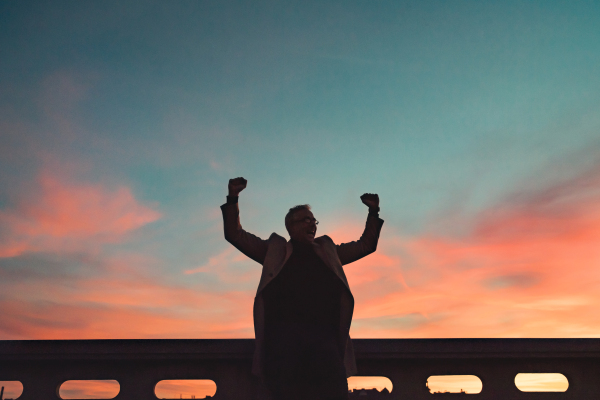 A silhouette of businessman standing on a bridge at dusk, expressing excitement by raising hands. Copy space.