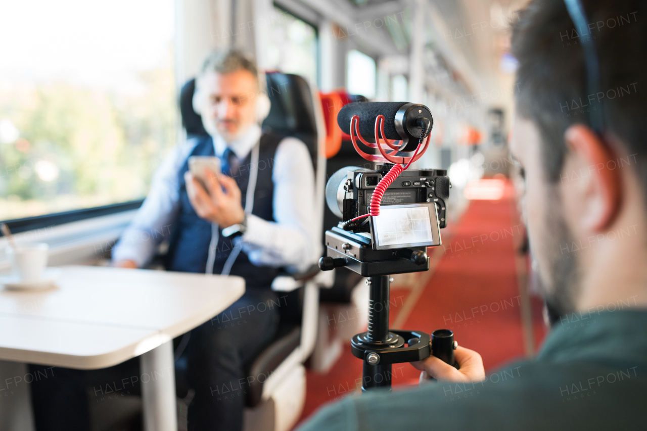 Unrecognizable man with video camera recording mature businessman with smartphone and headphones travelling by train.