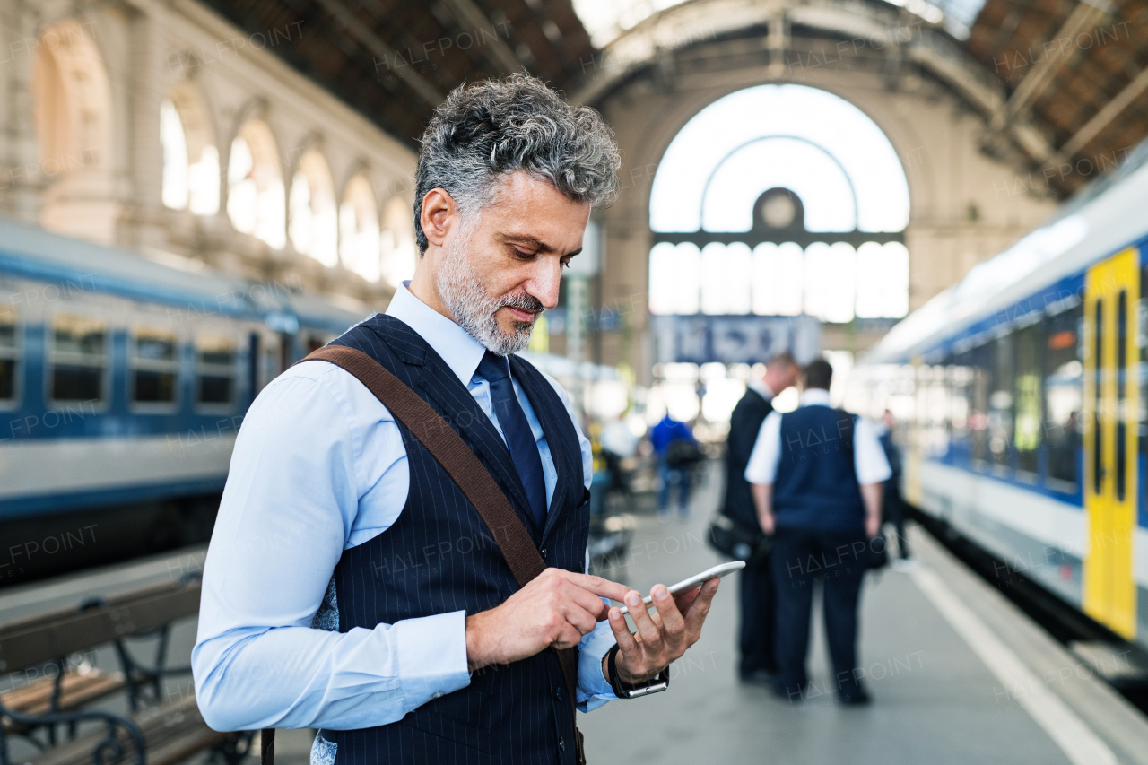 Handsome mature businessman with smartphone in a city. Man waiting for the train at the railway station, text messaging.