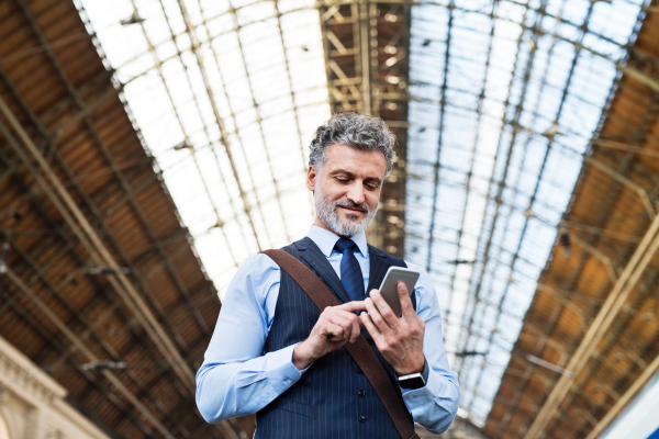 Handsome mature businessman with smartphone in a city. Man waiting for the train at the railway station, text messaging.