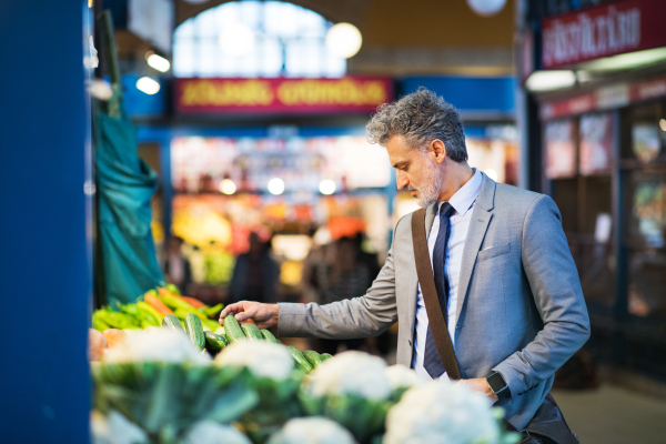 Handsome mature businessman in a city, buying vegetables from a vendor. Blurred background.