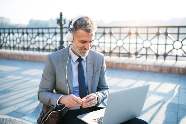 Handsome mature businessman with laptop in a city. Man sitting on a bridge, using laptop and headphones.