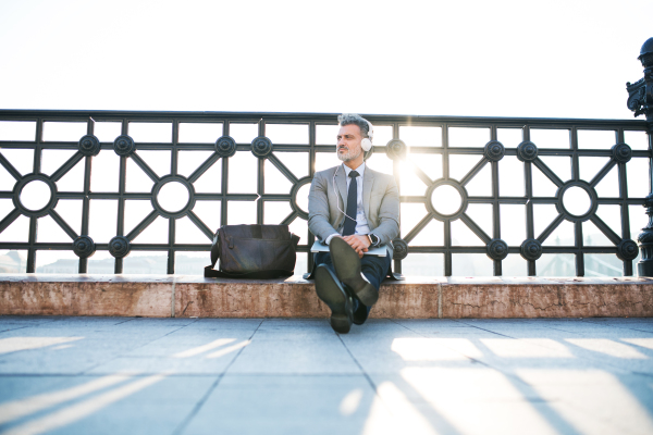 Handsome mature businessman with laptop in a city. Man sitting on a bridge, using headphones.