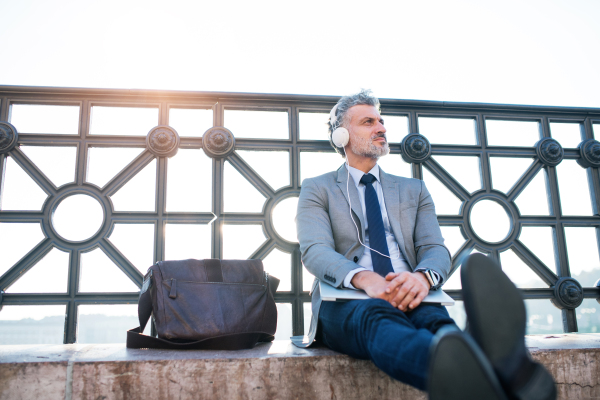 Handsome mature businessman with laptop in a city. Man sitting on a bridge, holding laptop and using headphones.