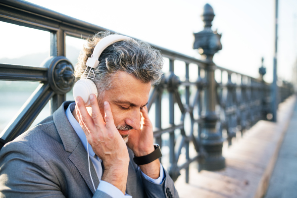 Handsome mature businessman with headphones in a city. Man sitting on a bridge, listening to music.