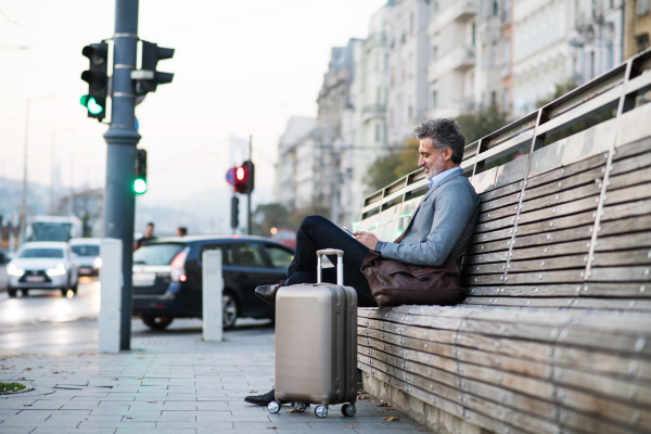 Handsome mature businessman with smartphone in a city, sitting on a bench, texting.