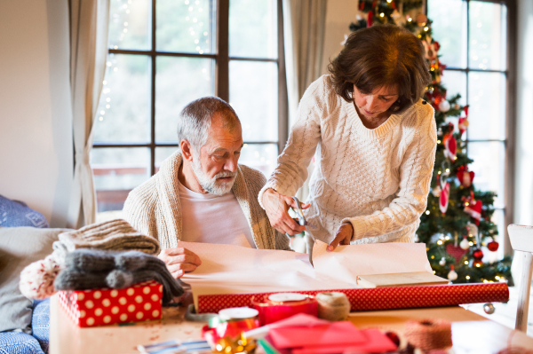 Beautiful senior couple in white woolen sweaters sitting at the table wrapping Christmas gifts together.