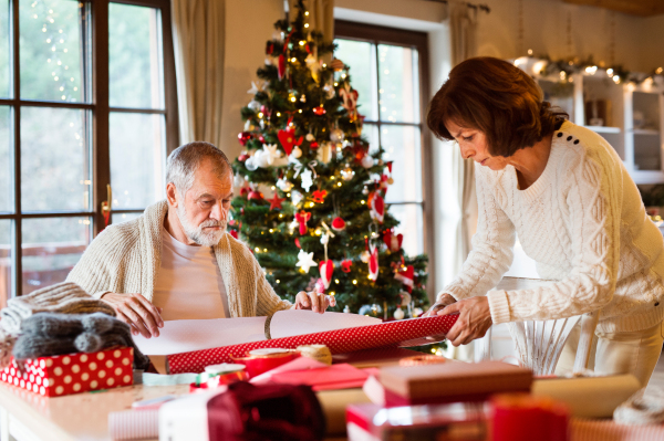 Beautiful senior couple in white woolen sweaters sitting at the table wrapping Christmas gifts together.